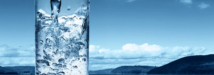 A tall glass of water being poured, set against a calm glacial lake and forested hills