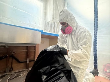 Worker in white Tyvek suit and respirator performing remediation under a kitchen sink in a containment zone
