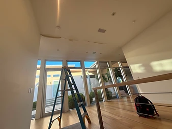 Modern interior room with light hardwood floors, floor-to-ceiling windows, blue Werner ladder, and long ceiling crack visible