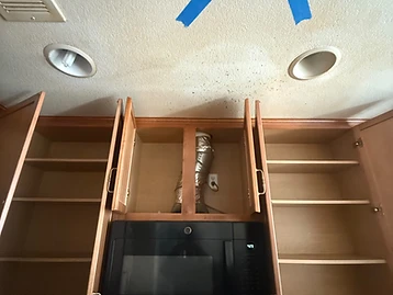 Close-up of kitchen cabinets and ceiling with blue painter's tape X marking water-stained area above microwave