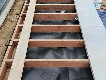 Wooden deck framing around an in-ground swimming pool with joists, landscape fabric, and partially installed stone tiles