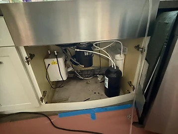 Kitchen cabinet under a stainless steel farmhouse sink showing garbage disposal, water filtration system, and instant hot water dispenser