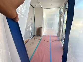 Hand opening a blue-zippered plastic containment barrier revealing a hallway under renovation with floor protection and plastic sheeting