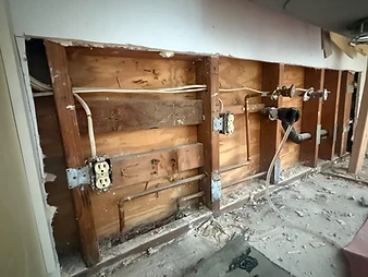 Exposed wall cavity under kitchen counter showing wood studs, electrical outlets, copper pipes, ABS drain, and debris on floor