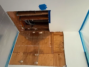 Ceiling with rectangular drywall section removed exposing joists and subfloor, plywood patch with moisture staining visible below