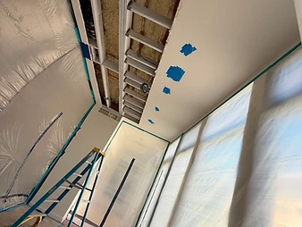 Low-angle view of open ceiling cavity with exposed joists, spray foam insulation, blue stepladder, and plastic containment walls