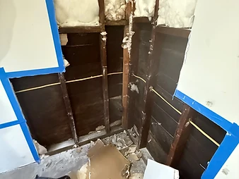 Wall cavity corner opened during remediation showing dark moisture-damaged studs, fiberglass insulation, and drywall debris on floor