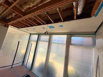 Room under containment with ceiling section removed, exposed joists, spray foam insulation, and plastic sheeting walls