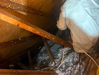 Worker in white hooded protective suit maneuvering in tight attic crawlspace among rafters and insulation