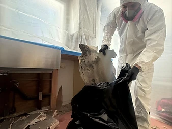 Worker in white Tyvek suit and respirator holding mold-covered drywall debris and placing it into a black trash bag
