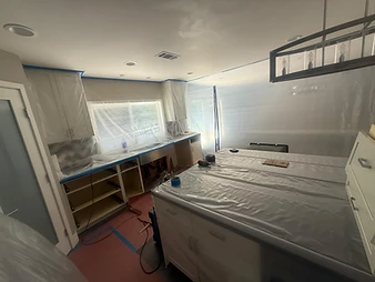 Kitchen prepped for containment with plastic sheeting covering cabinets and island, cordless drill on counter, and floor protection paper