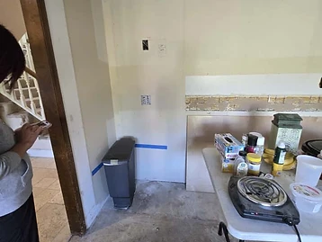 Kitchen area under repair with folding table holding supplies, exposed drywall framing, and open ceiling joists