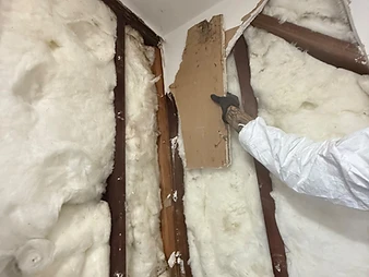 Worker in white Tyvek suit and gloves pulling drywall debris from wall cavity, tattoos visible on forearm