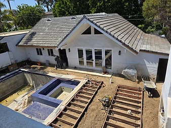 Aerial view of residential construction site with unfinished pool, wooden deck framing, yellow compressor, wheelbarrow, and white house extension