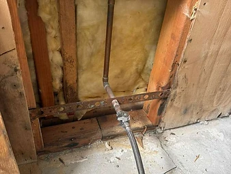 Close-up of exposed wall cavity with copper piping, angle stop valve, braided supply line, and Sioux Chief toilet floor flange on concrete subfloor
