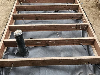 Rectangular wooden deck framing on bare earth with black landscape fabric, PVC pipes, and electrical wiring through joists