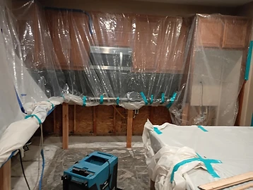 Kitchen workspace enclosed in plastic sheeting with blue painter's tape, XPOWER dehumidifier, and covered island