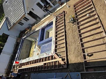 Aerial view of backyard construction with unfinished pool, wooden deck framing, yellow air compressor, and house with French doors