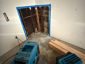 Exposed wall cavity with dark stained studs, blue painter's tape border, and two XPOWER air movers on the floor