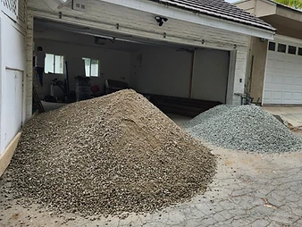 Exterior of white brick house with gravel piles, cracked asphalt driveway, and palm trees visible