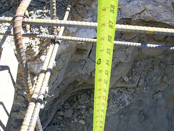 Yellow tape measure held against vertical rebar and excavated earth trench wall showing foundation depth measurement