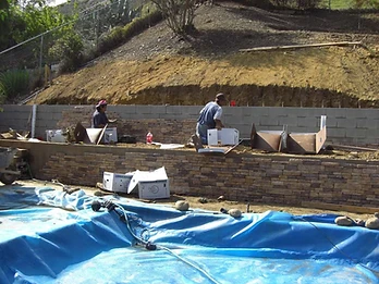 Two workers installing stone veneer on a concrete block retaining wall with Silverado Stone boxes, blue pool cover in foreground