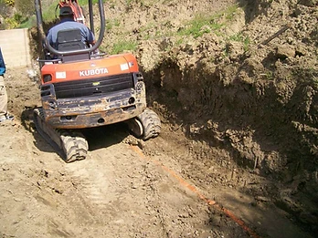 Orange Kubota mini-excavator digging a foundation trench with orange drainage pipe and partial CMU block wall visible