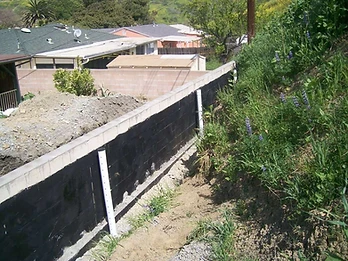 Retaining wall with black waterproofing coating and two white PVC drainage pipes against a grassy embankment