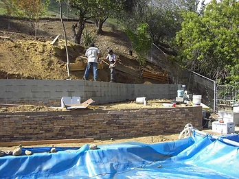 Two-tier retaining wall with stone veneer and concrete block upper wall, workers leveling soil behind, blue pool cover in foreground