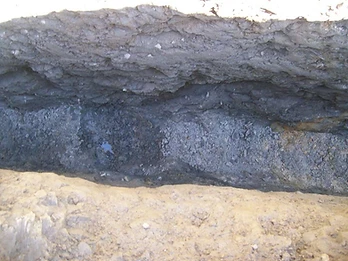 Close-up of an open foundation trench showing compacted earth, standing water, and gravel drainage bedding at the base