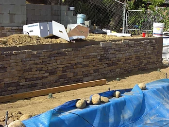 Partially completed stone veneer retaining wall with Silverado Stone box, blue pool cover, drainage caps, and dirt construction area