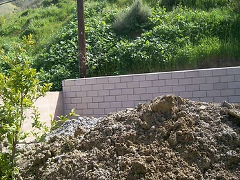 Newly constructed concrete block retaining wall with excavated soil pile and grassy hillside behind