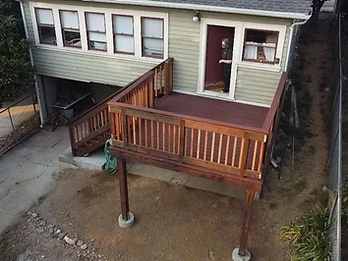 Elevated wooden deck with dark reddish-brown stain and natural wood balusters attached to a sage-green house with concrete pier footings