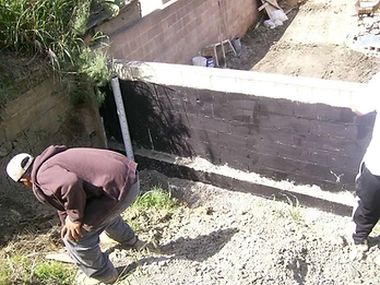 Retaining wall with black waterproofing coating and drainage pipe installed, worker crouched nearby