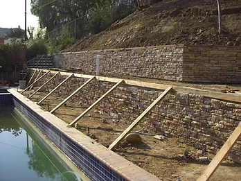 Two-tier stone veneer retaining wall with diagonal wooden bracing, blue pool cover, and PVC drainage pipe between tiers