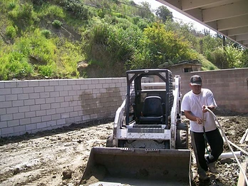 Worker holding a pipe near a white skid-steer loader behind a newly built concrete block retaining wall