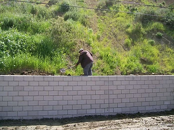 Worker using a shovel near a partially completed concrete block retaining wall on a hillside