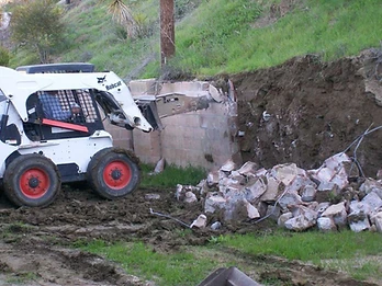 White skid-steer loader demolishing a concrete masonry block wall with rubble accumulating on a muddy construction site