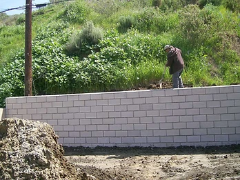 Worker using a shovel to level soil behind a newly constructed concrete block retaining wall with utility pole nearby