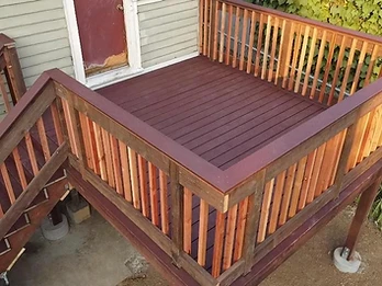 Elevated wooden deck with dark burgundy stain, natural amber balusters, and staircase on concrete pier footings against sage-green house siding