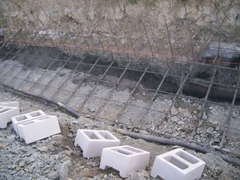 Concrete masonry blocks staged along a trench edge with rebar grid and black drainage pipe installed below