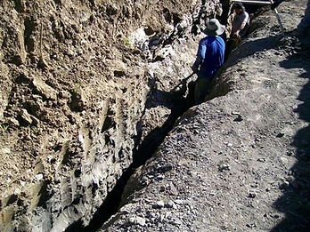 Two workers standing alongside a deep narrow foundation trench with exposed earth walls on a sunny construction site