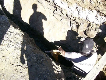 Worker in a Dodgers cap operating a yellow jackhammer to break rock at the base of a narrow foundation trench