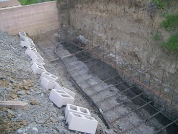 Concrete masonry blocks staged along excavation edge with rebar grid and corrugated drainage pipe in trench below
