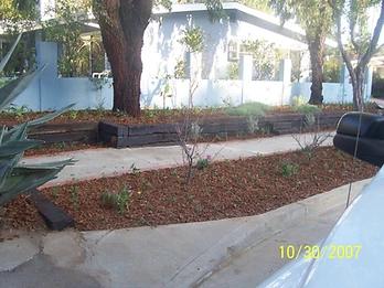 Completed landscaping with concrete walkway, mulched garden bed, low timber retaining wall, and periwinkle-blue house