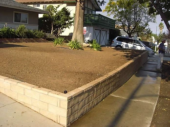 Completed L-shaped tan concrete block retaining wall with leveled garden bed, trees, and concrete sidewalk in a residential yard