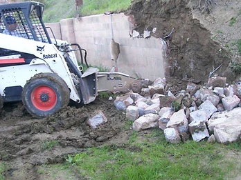 White skid-steer loader with hydraulic breaker demolishing an existing concrete block retaining wall with debris falling