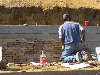Worker kneeling and installing decorative stone veneer on a concrete block retaining wall with rebar studs exposed at top
