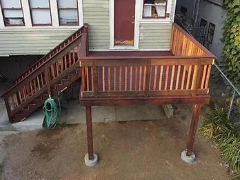 High-angle view of completed elevated wooden deck with dark maroon stain, natural wood balusters, and staircase descending to concrete patio