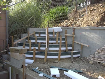 Wooden and foam-board formwork erected against a cinder block wall with green planks and white foam segments on a dirt construction site